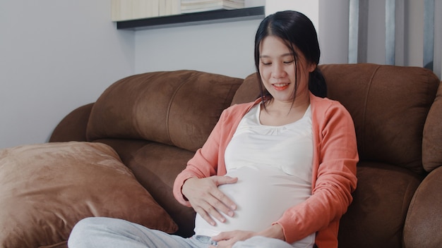 Young Asian Pregnant woman holding her belly talking with her child. Mom feeling happy smiling positive and peaceful while take care baby, pregnancy lying on sofa in living room at home .